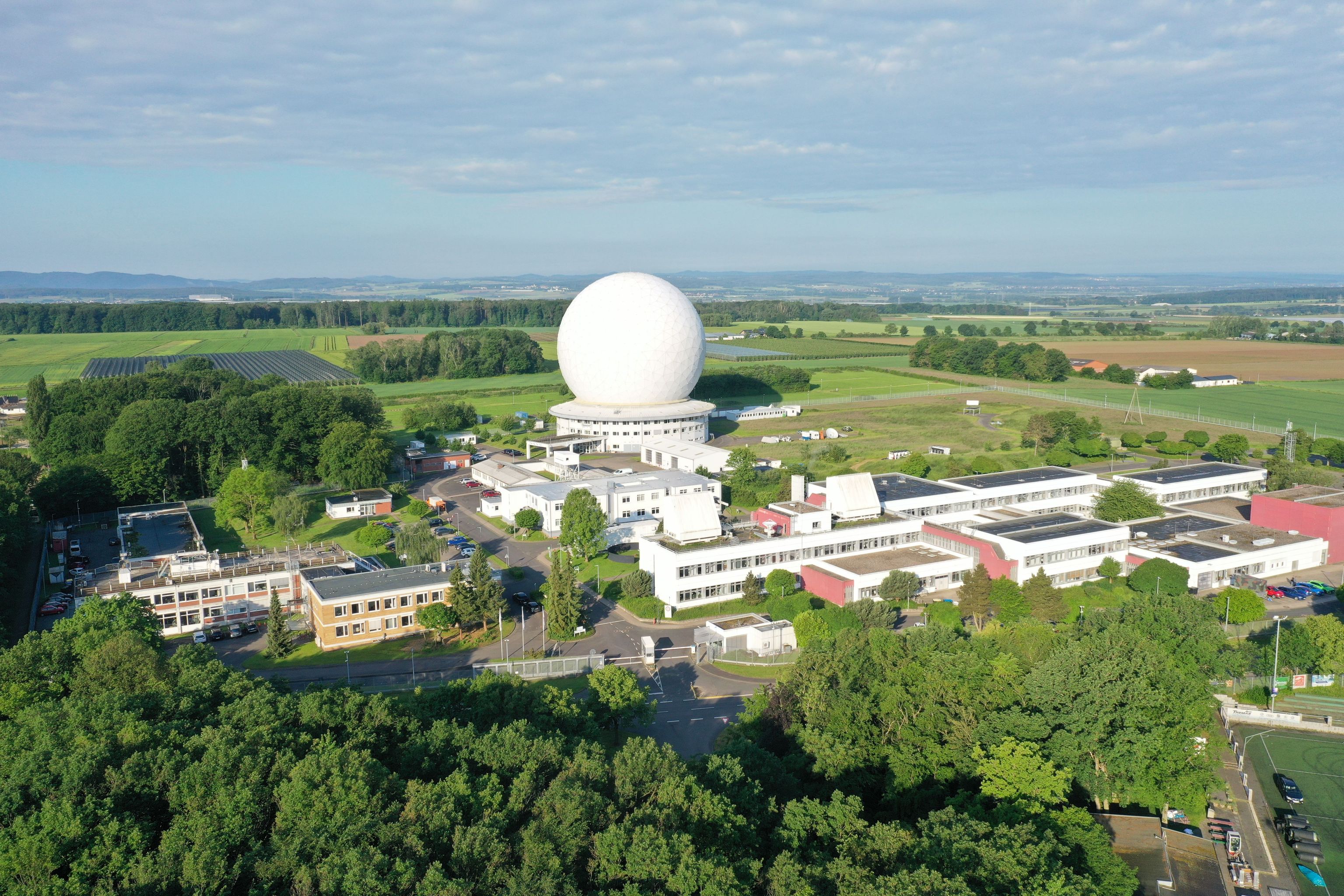 Aerial view: Großes Gebäude mit markanter Kuppel und grünem Umfeld, umgeben von Feldern und Wald.
