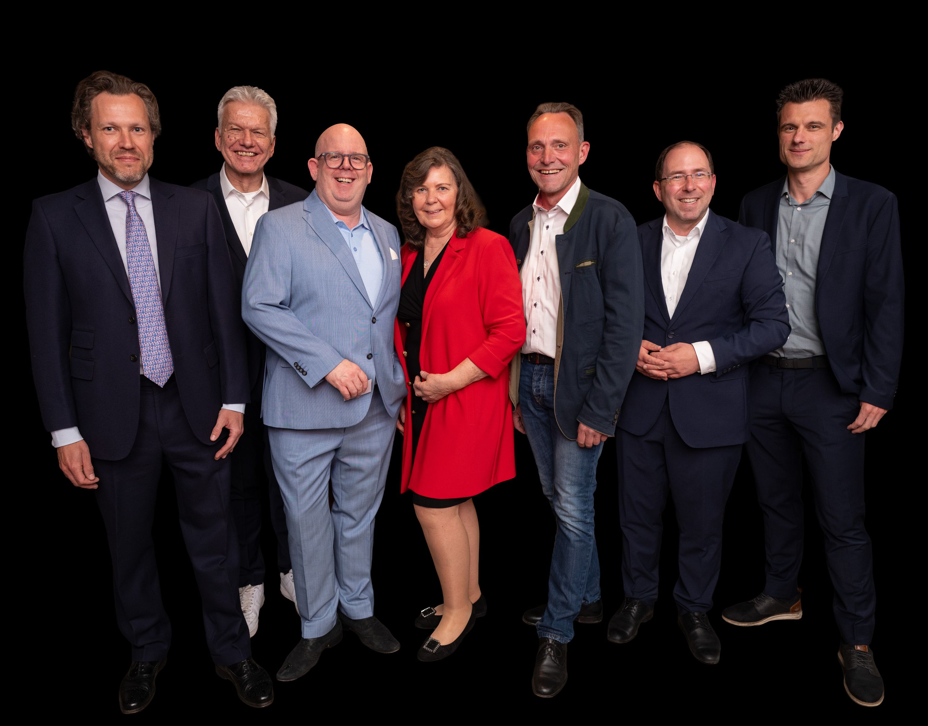 Sechs Personen in Anzügen und eine Frau im roten Kleid stehen lächelnd für Gruppenfoto.