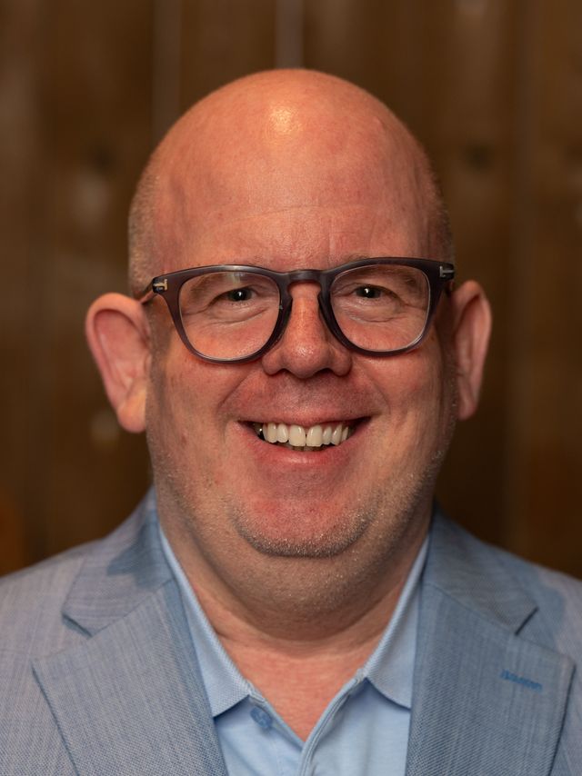 Lächelnder Mann mit Brille in blauem Hemd und grauer Jacke.