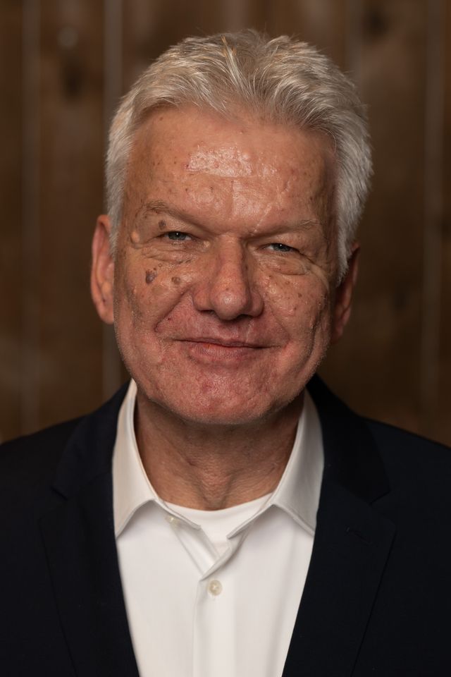 Älterer Herr mit freundlichem Lächeln, dunklem Blazer und weißem Hemd vor neutralem Hintergrund.