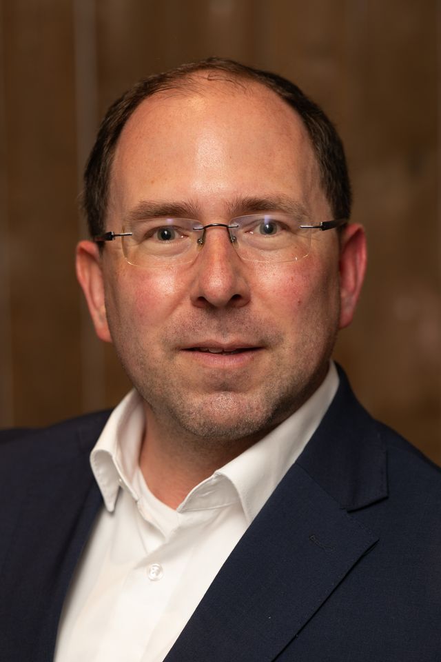 Porträt eines lächelnden Mannes mit Brille in dunkelblauem Sakko und weißem Hemd.