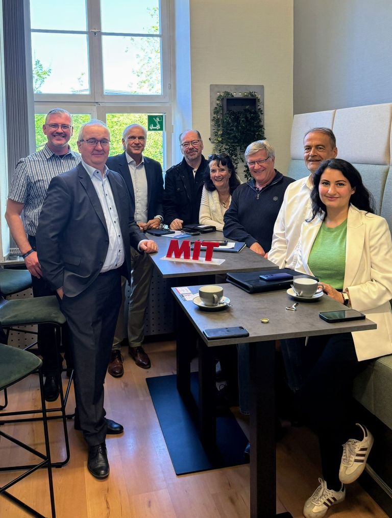 Gruppe von acht Personen bei einer Tischbesprechung mit Kaffeetablett und Namensschild „MIT“.