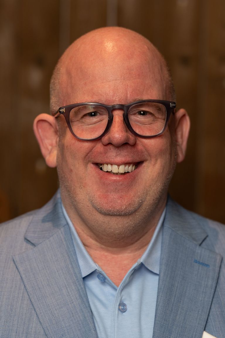 Lächelnder Mann mit Brille in blauem Hemd und grauer Weste.