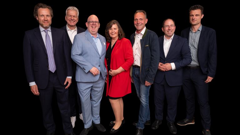 Sechs Personen in Anzügen und eine Frau im roten Kleid lächeln auf Gruppenfoto.