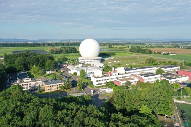 Aerial view: Großes Gebäude mit markanter Kuppel und grünem Umfeld, umgeben von Feldern und Wald.