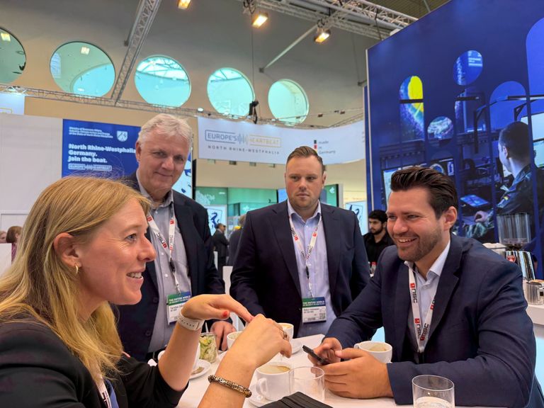 Vier Geschäftsleute in Diskussion auf einer Messe mit Informationsdisplay im Hintergrund.
