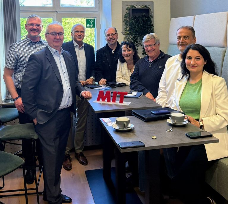Gruppe von acht Personen bei einer Besprechung an einem Tisch mit „MITT“-Schild.